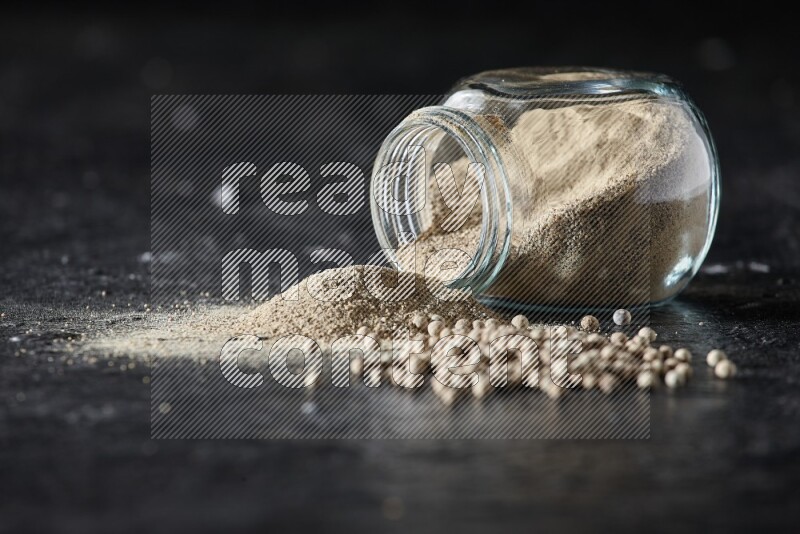A flipped herbal glass jar full of white pepper powder with spilled powder and pepper beads on textured black flooring