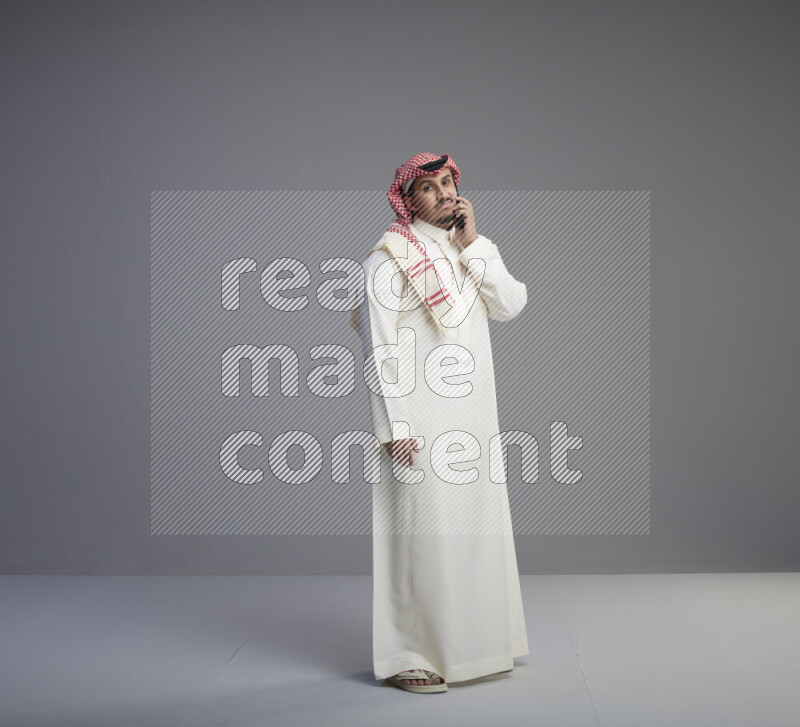 A Saudi man standing wearing thob and red shomag talking on phone on gray background