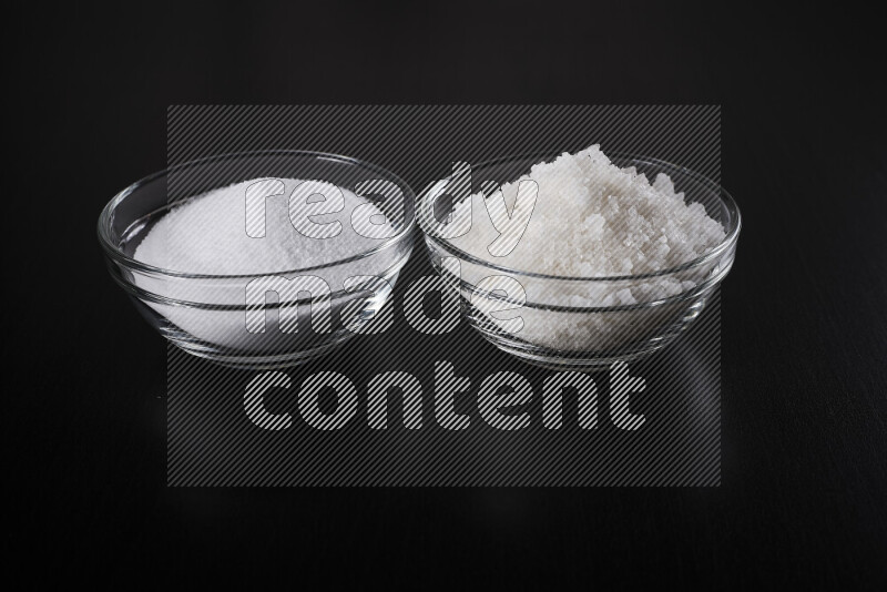 2 glass bowls one is filled with table salt and the other with coarse sea salt on black background