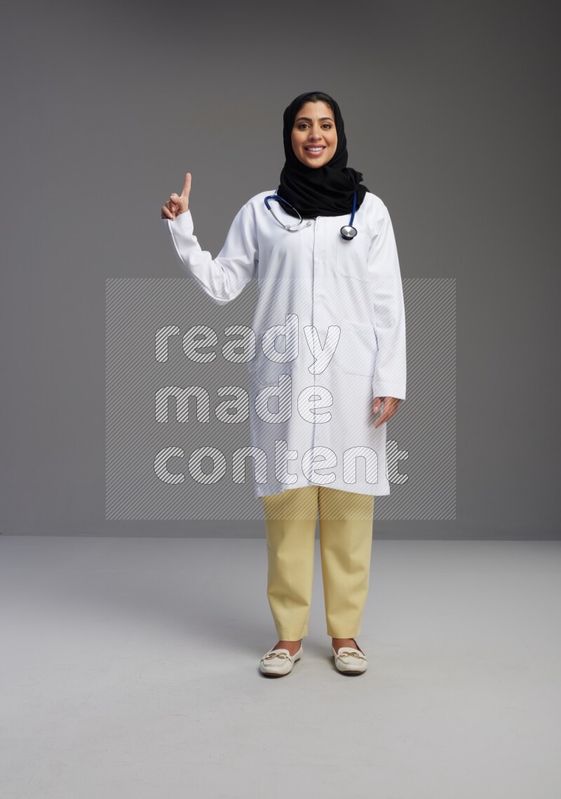 Saudi woman wearing lab coat with stethoscope standing interacting with the camera on Gray background