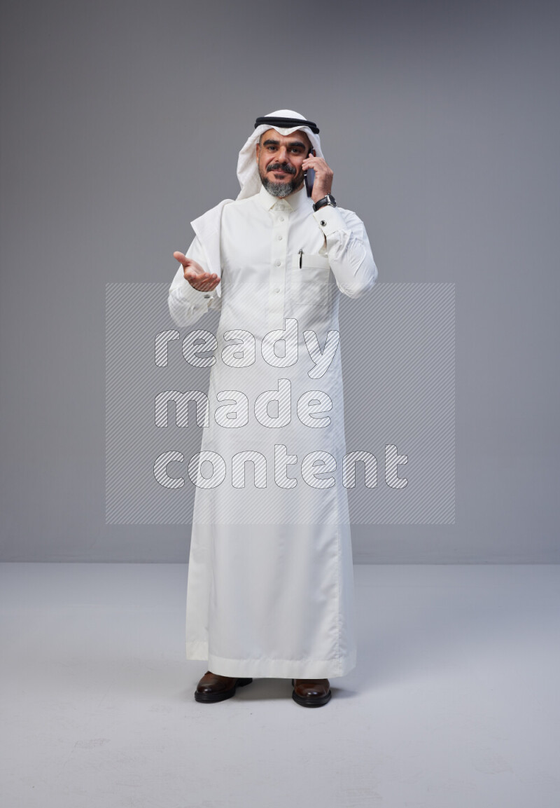Saudi man Wearing Thob and white Shomag standing talking on phone on Gray background