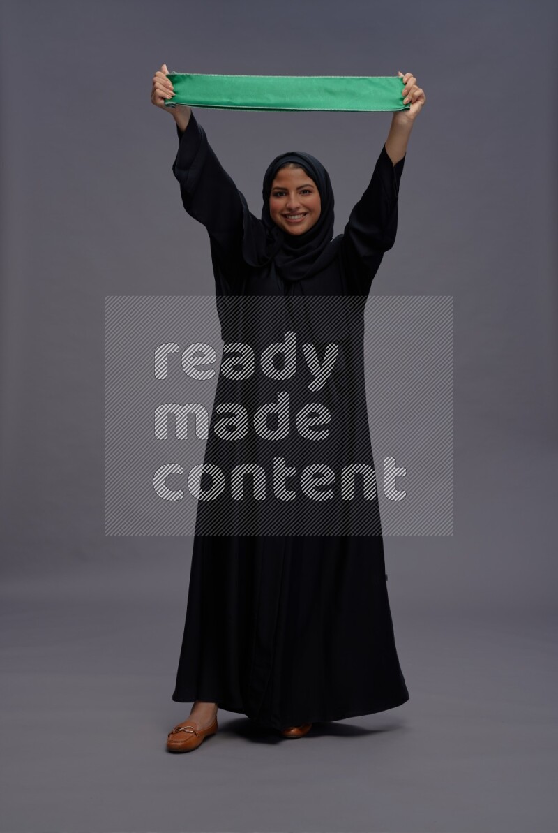 Saudi woman wearing Abaya standing holding Saudi flag on gray background