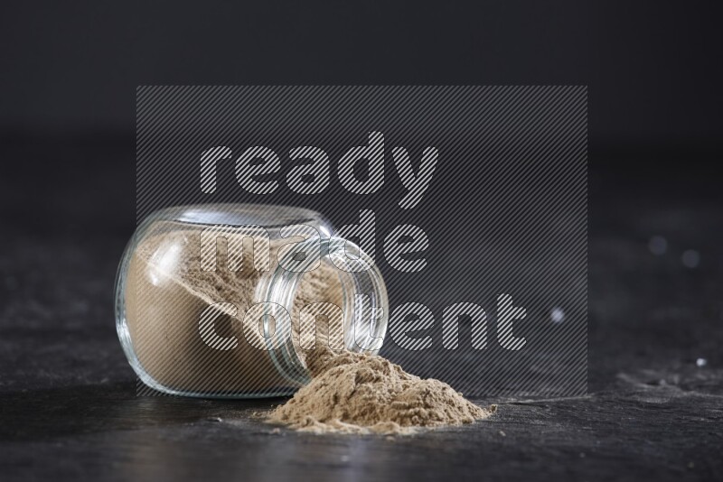 A glass spice jar full of garlic powder flipped over with the powder came out on a textured black flooring