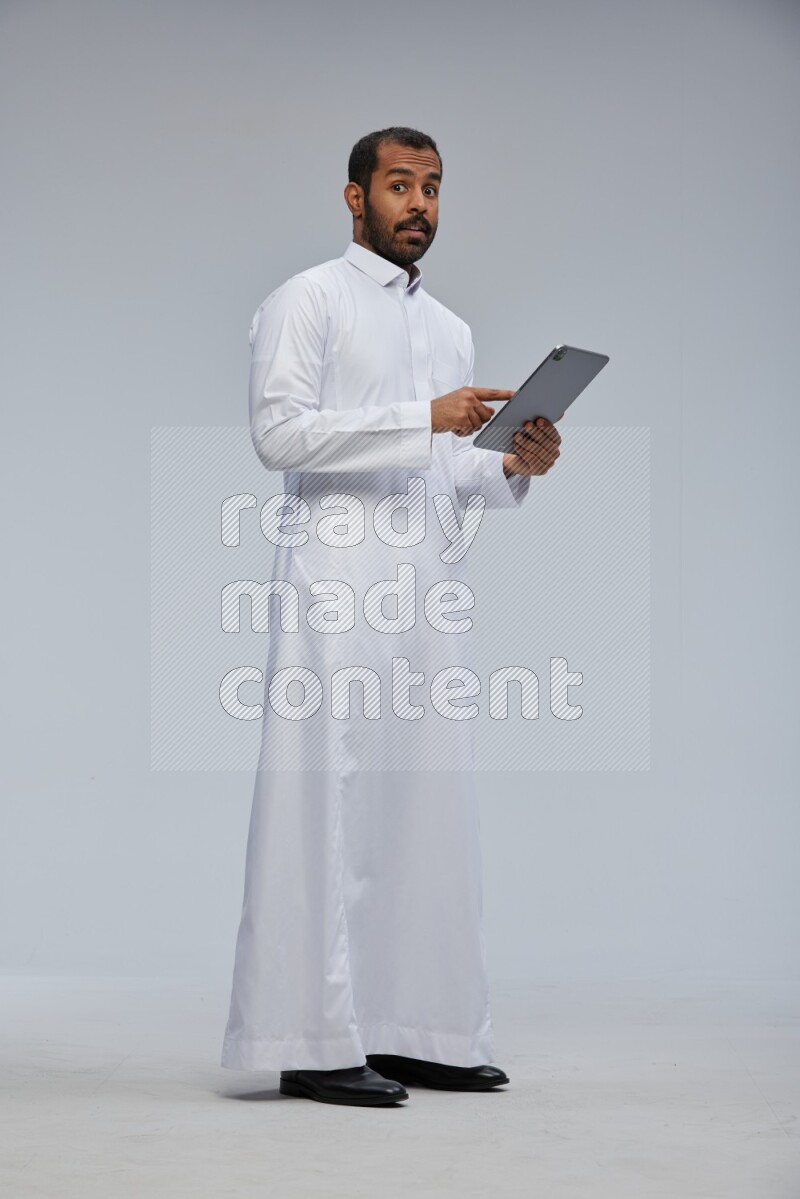 Saudi man wearing Thob standing working on tablet on Gray background