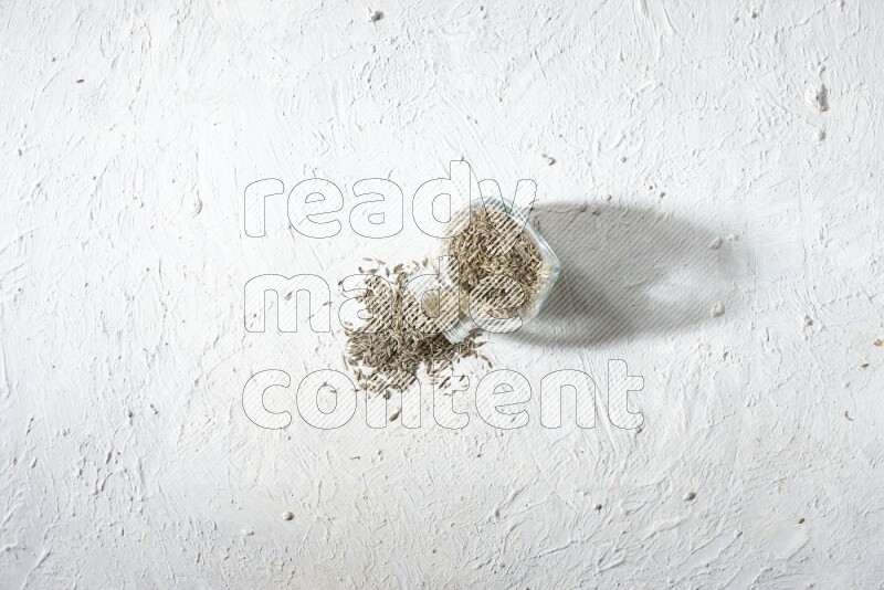 A flipped glass spice jar full of cumin seeds and the seeds spilled out on textured white flooring