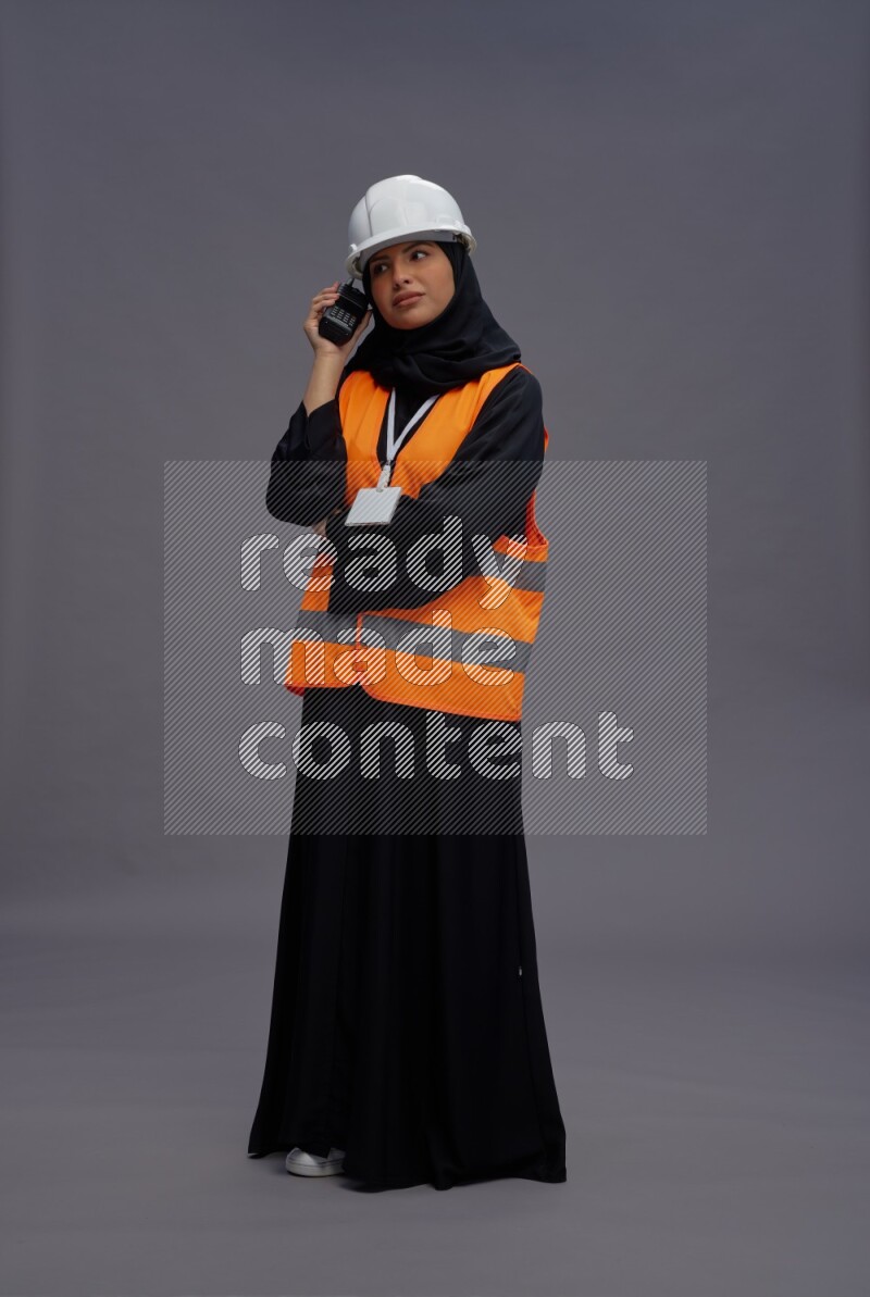 Saudi woman wearing Abaya with engineer vest with neck strap employee badge standing holding walkie-talkie on gray background