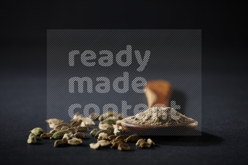 A wooden ladle full of cardamom powder and cardamom seeds beside it on black flooring