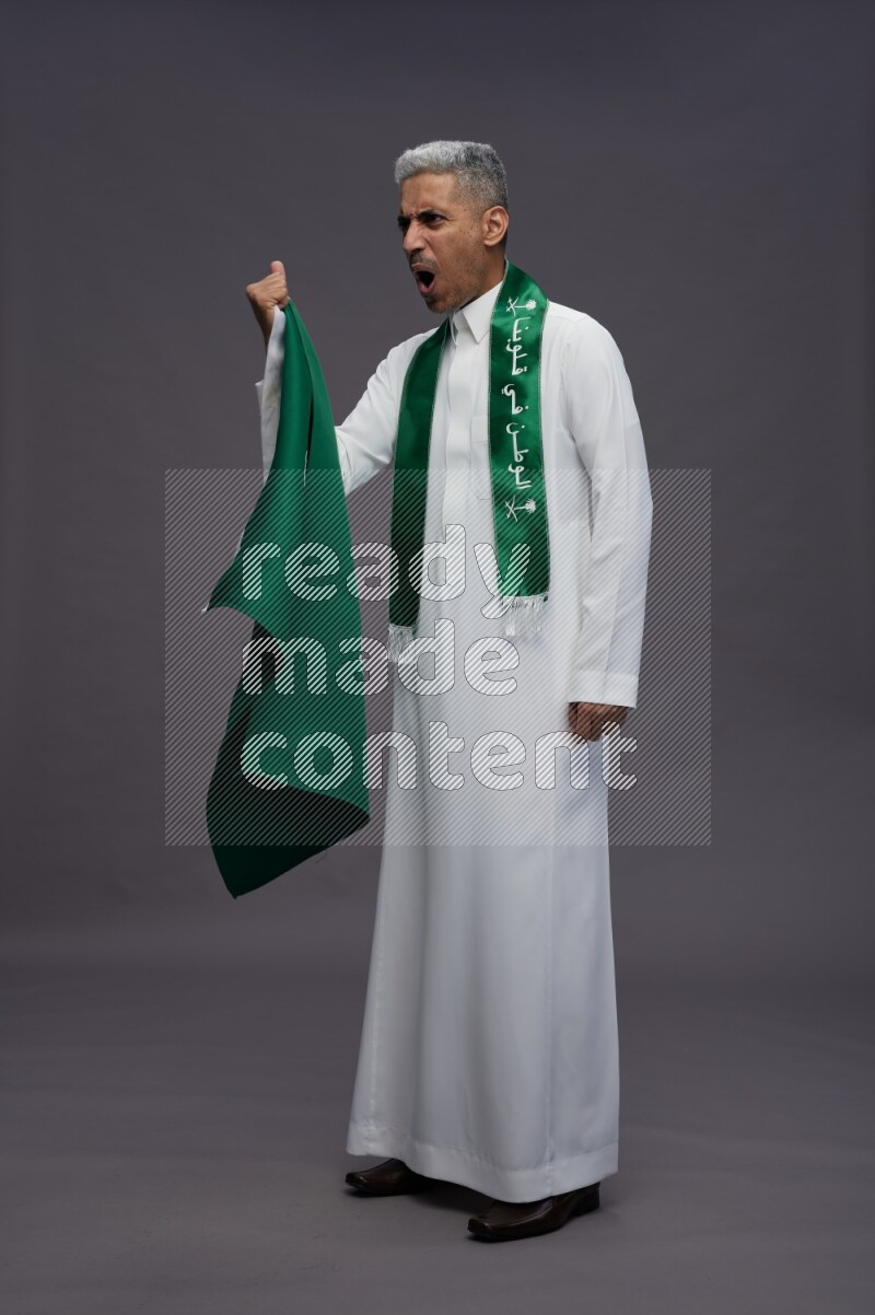 Saudi man wearing thob standing holding Saudi flag on gray background