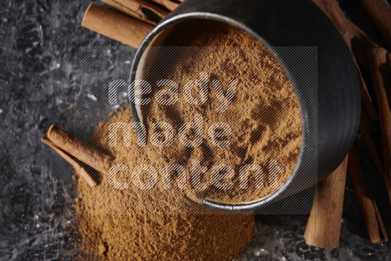 Black pottery bowl over filled with cinnamon powder and cinnamon sticks around the bowl on a textured black background