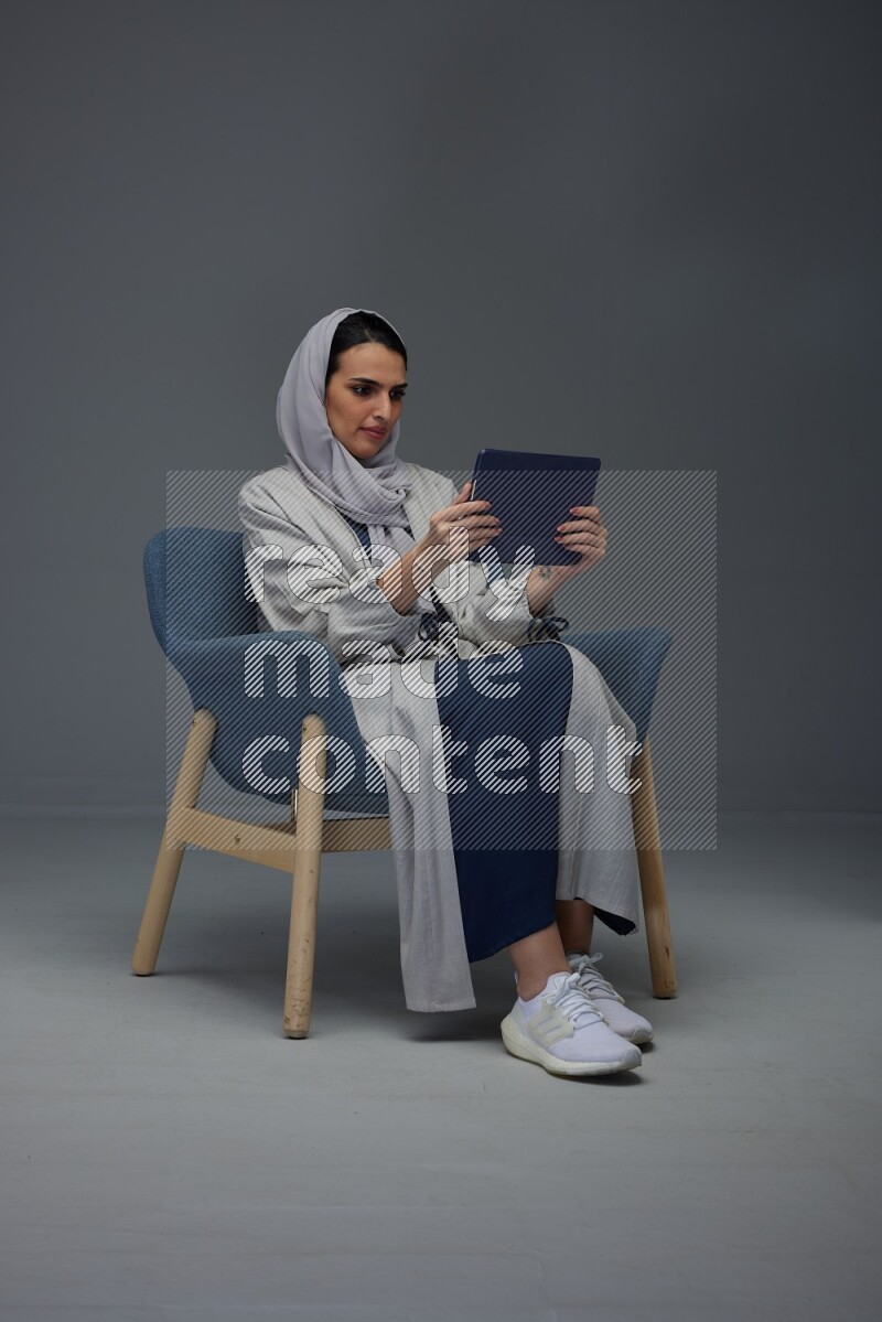 A Saudi female wearing a light gray Abaya and head scarf sitting on a dark grey chair while holding her iPad on a grey background