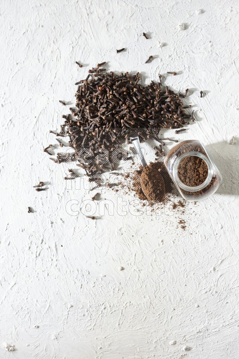 A glass spice jar and a metal spoon full of cloves powder and cloves spread on textured white flooring