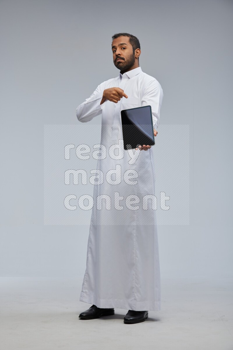 Saudi man wearing Thob standing showing tablet to camera on Gray background