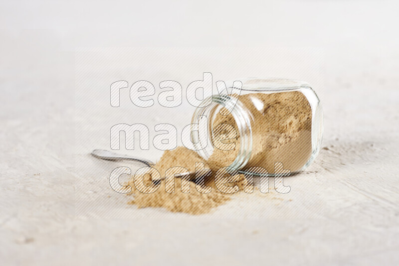 A glass jar full of ground ginger powder flipped with some spilling powder on white background