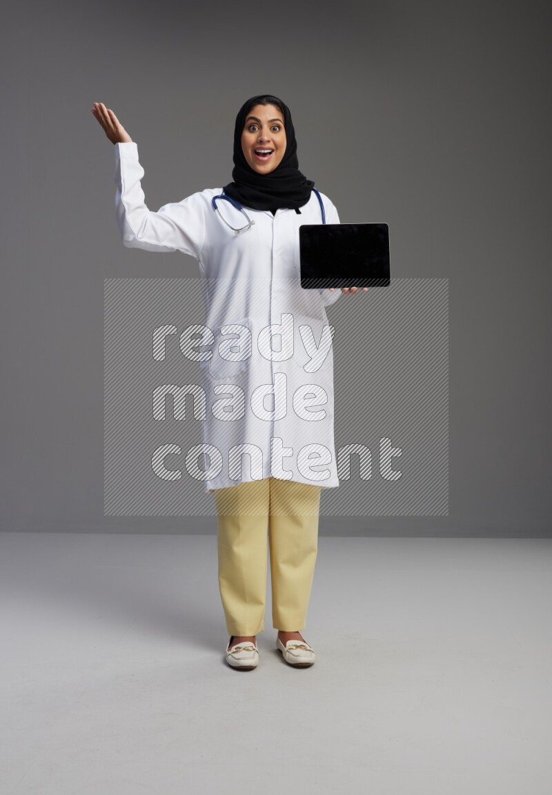 Saudi woman wearing lab coat with stethoscope standing showing tablet to camera with sign in the back on Gray background