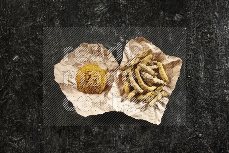 Turmeric powder and dried turmeric whole fingers in 2 crumpled pieces of paper on textured black flooring