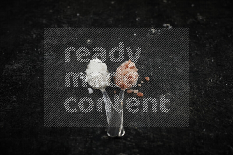 2 metal spoons filled with white salt and pink himalayan salt on black background