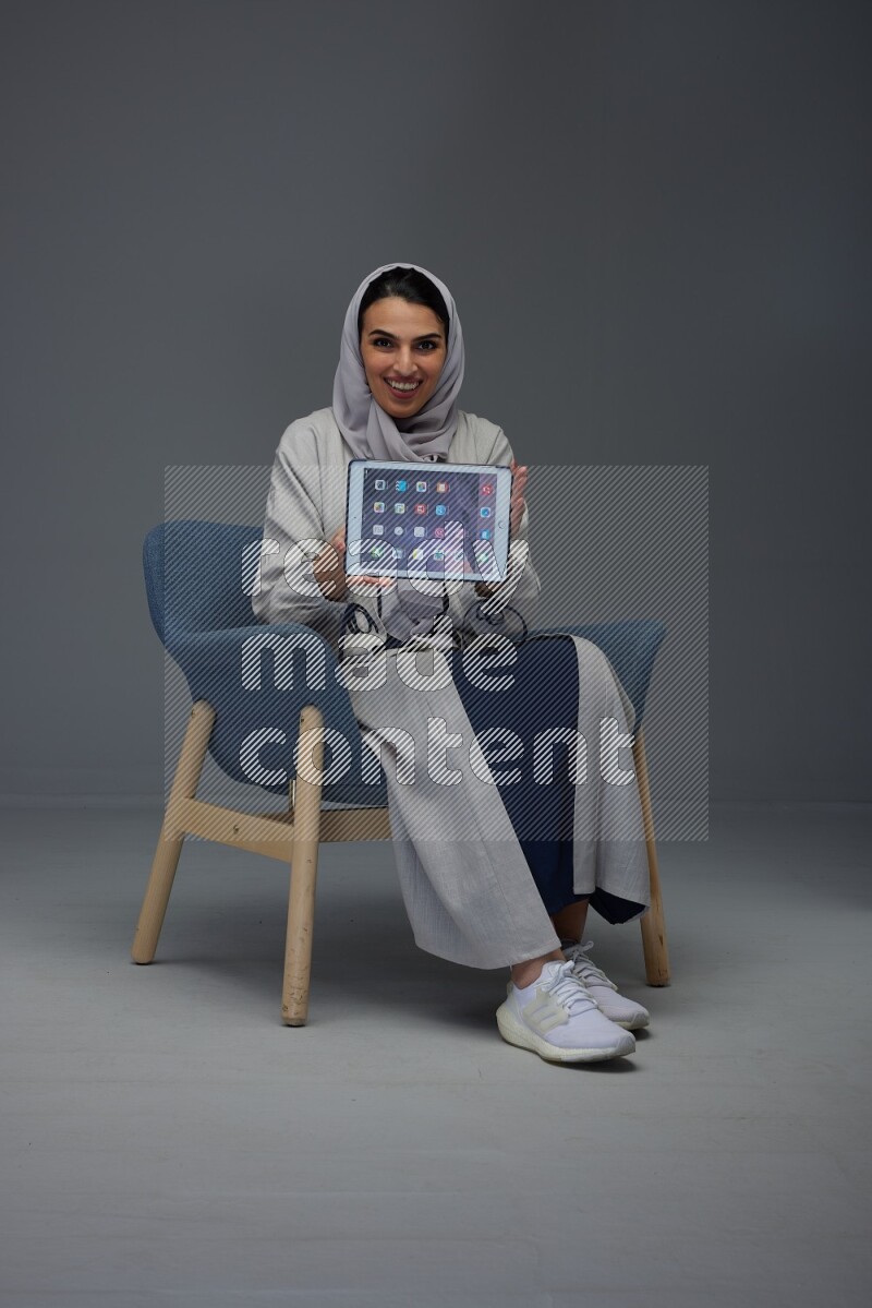 A Saudi woman wearing a light gray Abaya and head scarf sitting on a dark grey chair and showing the tablet's screen eye level on a grey background