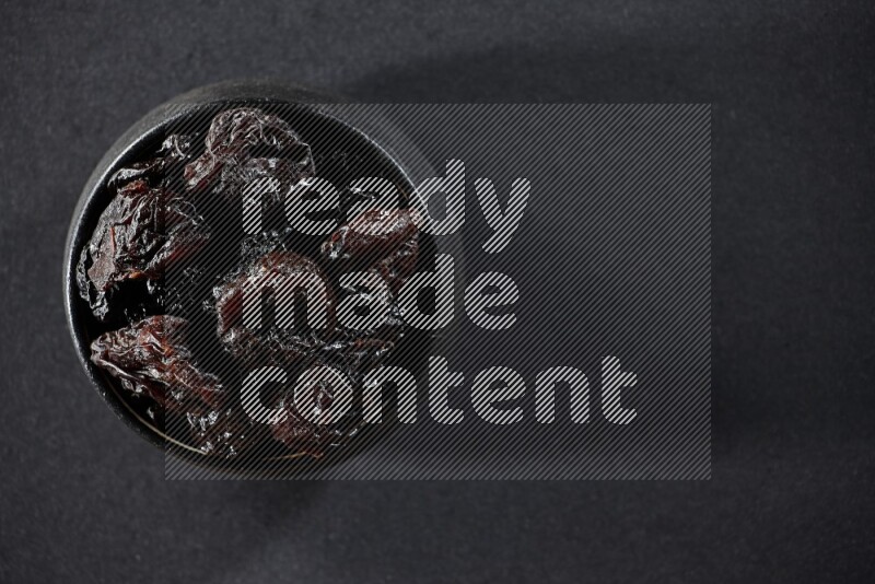 A black pottery bowl full of dried plums on a black background in different angles