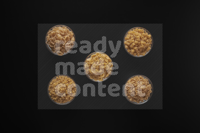 Different pasta types in 5 glass bowls on black background