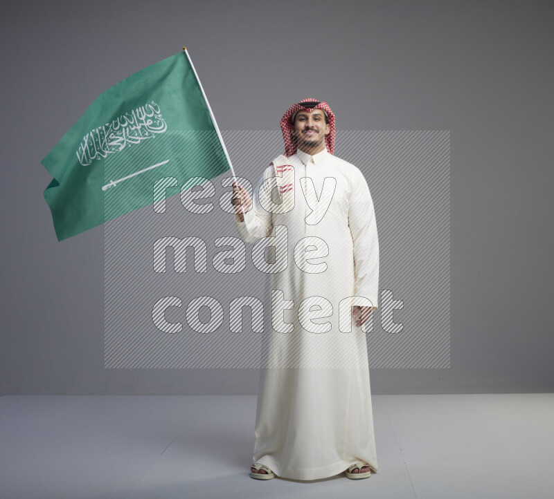 A Saudi man standing wearing thob and red shomag raising big Saudi flag on gray background