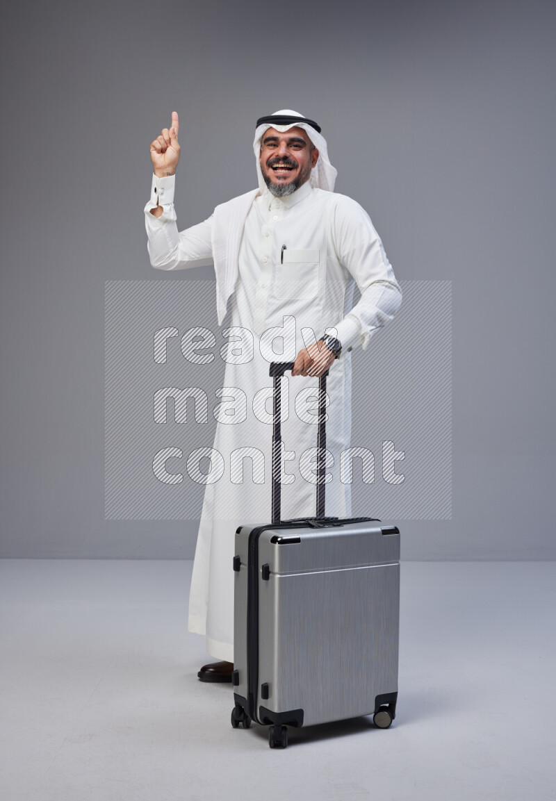 Saudi man wearing Thob and white Shomag standing holding Travel bag on Gray background