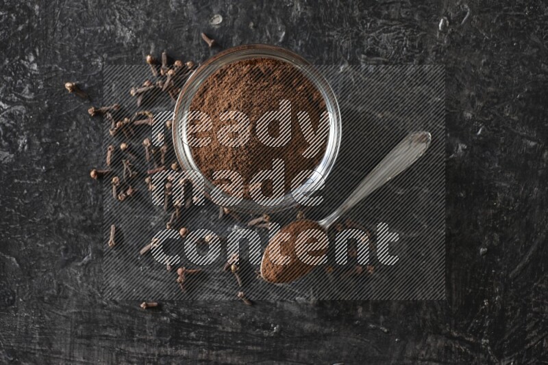 وعاء زجاجي وملعقة معدنية ممتلئان ببودرة القرنفل مع حبوب القرنفل بجانبهما على خلفية سوداء