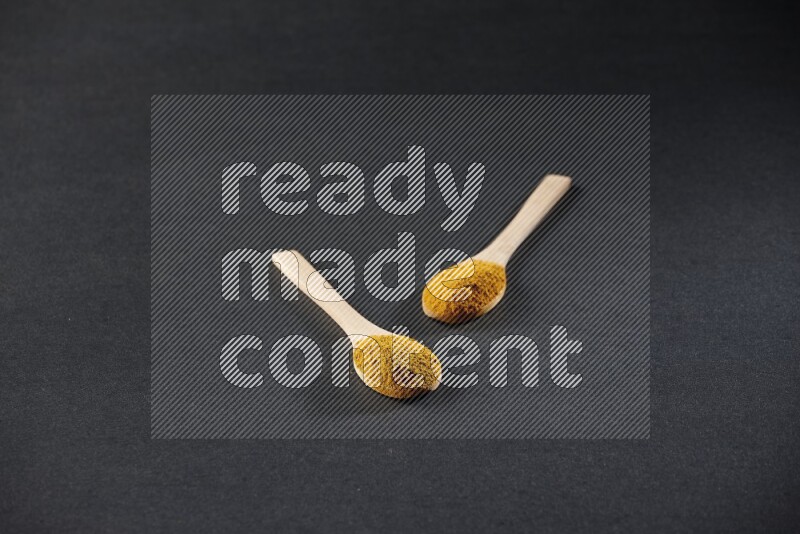 2 wooden spoons full of turmeric powder on black flooring