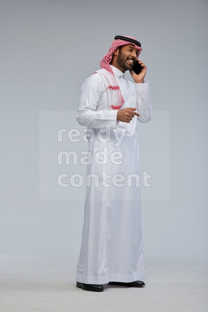 Saudi man Wearing Thob and shomag standing talking on phone on Gray background