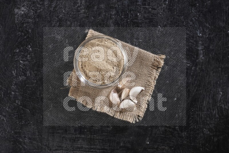 A glass bowl full of garlic powder placed on burlap fabric with garlic cloves on a textured black flooring