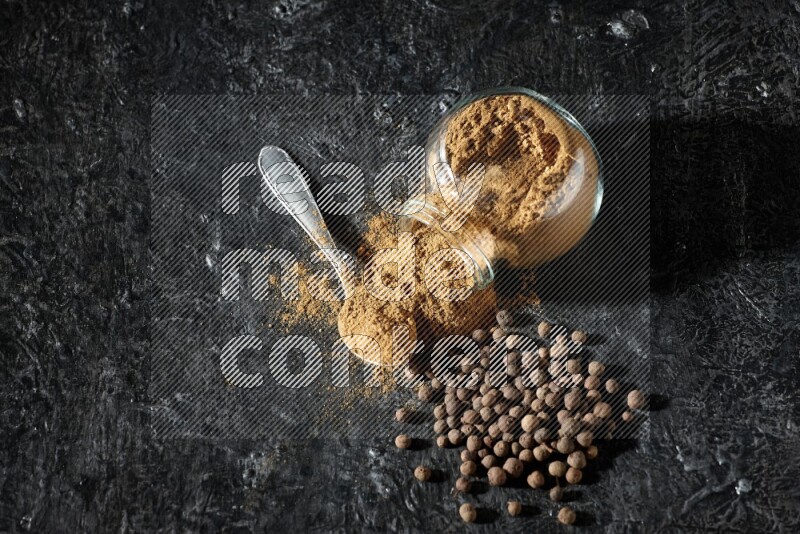 وعاء زجاجي للتوابل مقلوب وملعقة معدنية ممتلئان ببودرة بهار الفلفل الحلو والبودرة تناثرت من الوعاء مع بعض الحبوب على خلفية سوداء