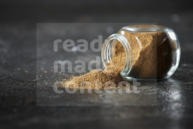 A flipped glass spice jar full of cumin powder and powder spilled out on a textured black flooring