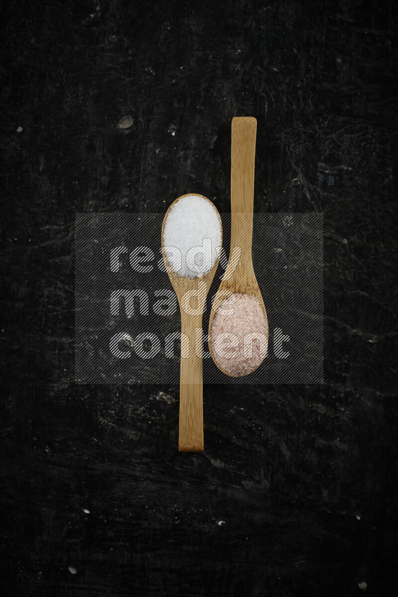 2 wooden spoons filled with fine and coarse salt on black background