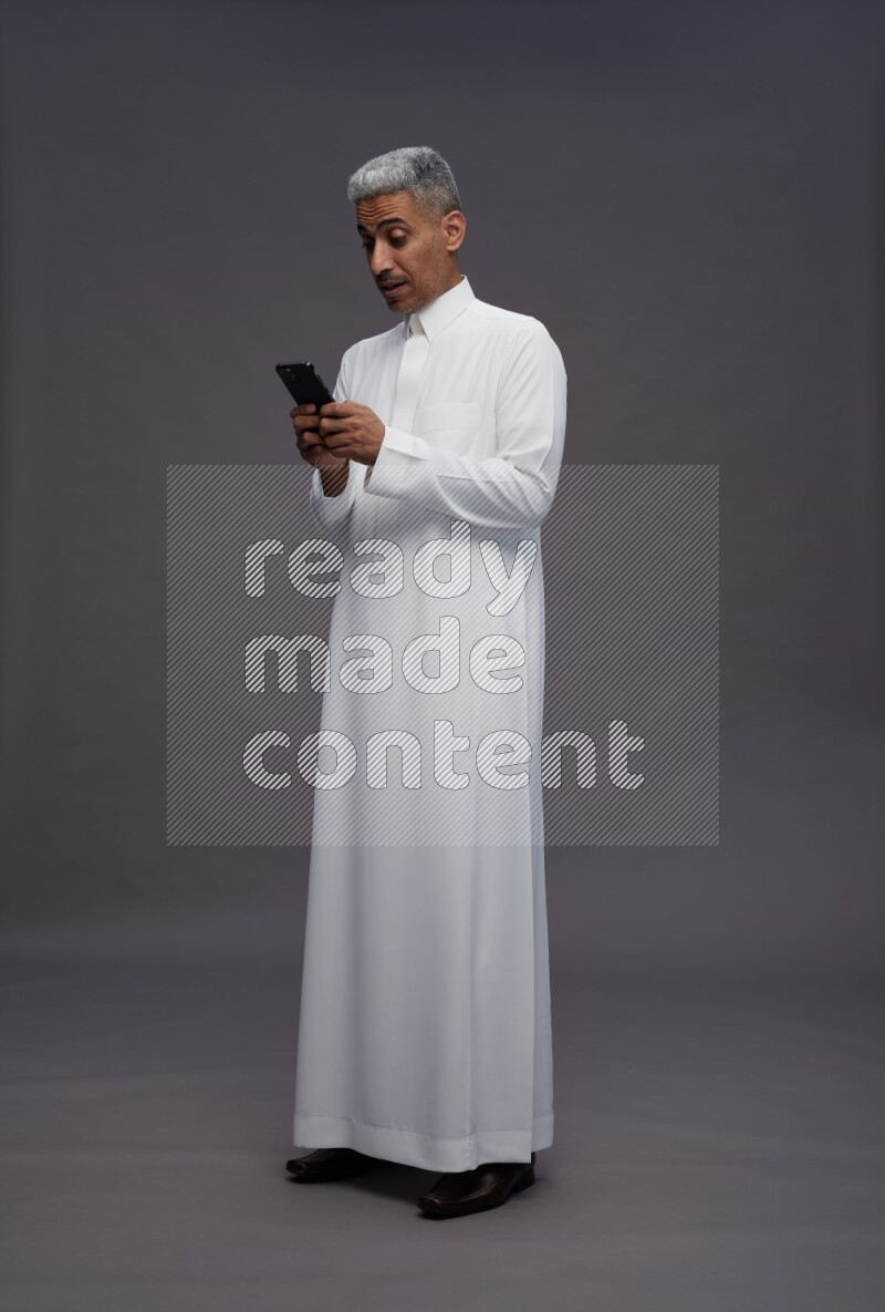 Saudi man wearing thob standing texting on phone on gray background