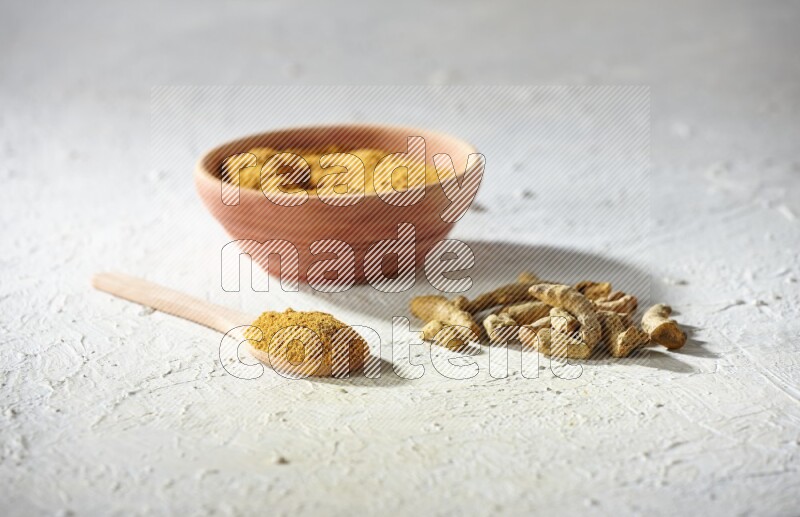A wooden bowl and wooden spoon full of turmeric powder with dried turmeric fingers on textured white flooring