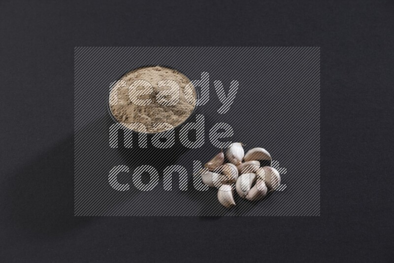 وعاء فخاري أسود ممتلئ ببودرة الثوم مع بعض فصوص الثوم بجانبه علي خلفية سوداء