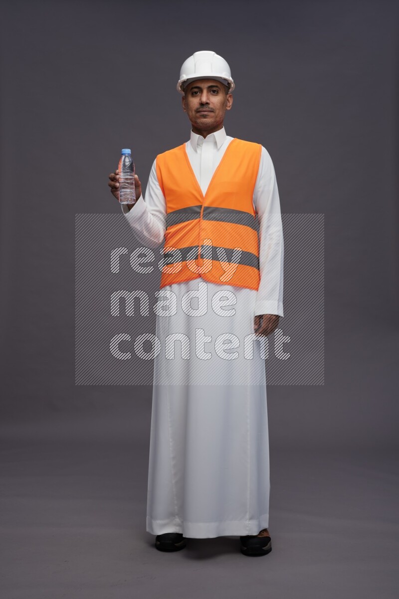Saudi man wearing thob with engineer vest standing drinking water on gray background