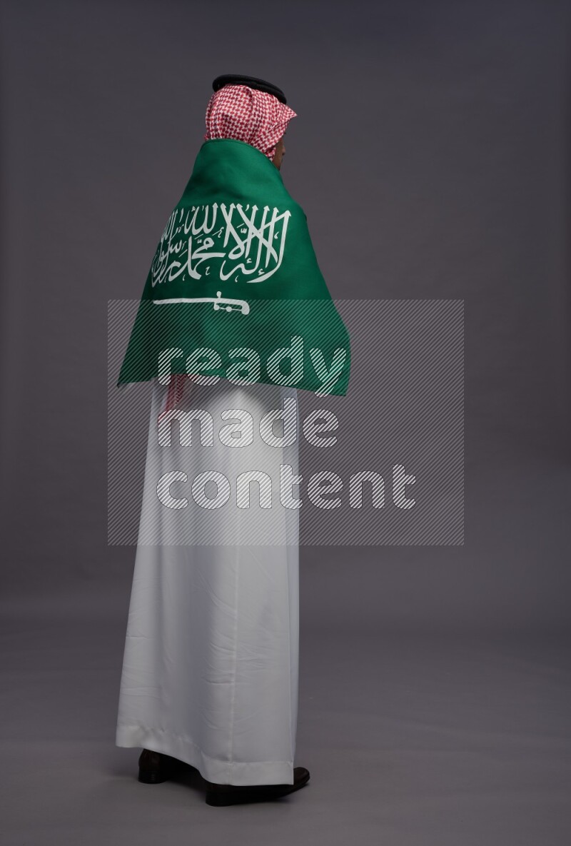 Saudi man wearing thob and shomag standing holding Saudi flag on gray background