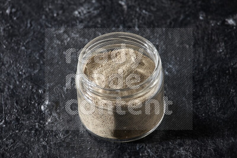 A glass jar full of white pepper powder on textured black flooring
