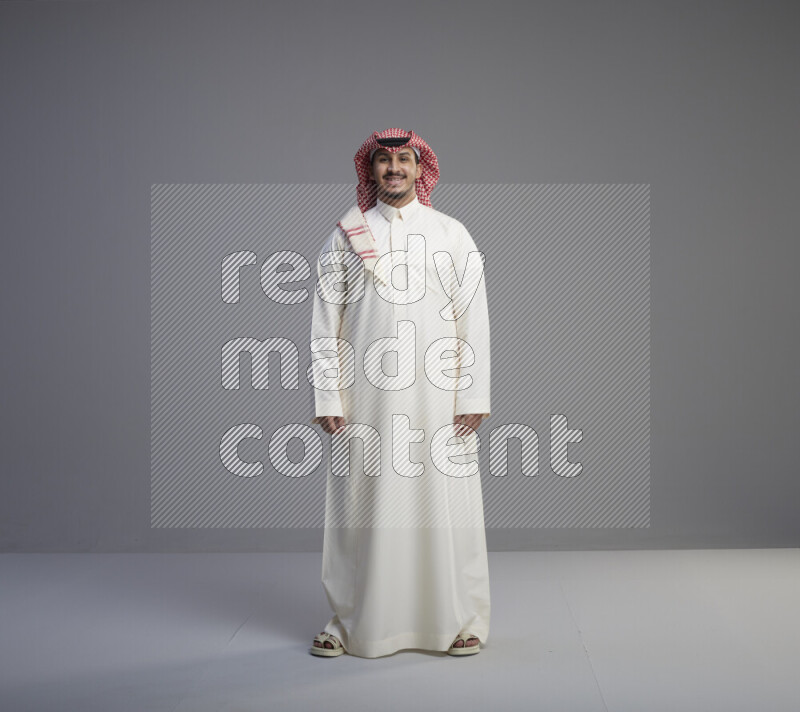 A Saudi man standing wearing thob and red shomag interacting with the camera on gray background