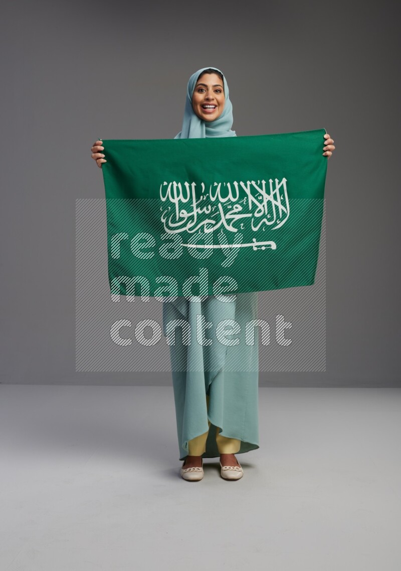 Saudi Woman wearing Abaya standing holding Saudi flag on Gray background