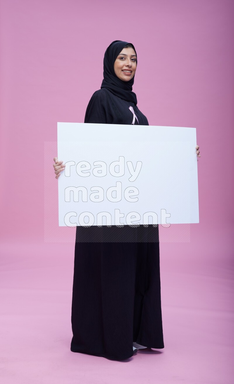 Saudi woman wearing pink ribbon on Abaya standing holding board on pink background
