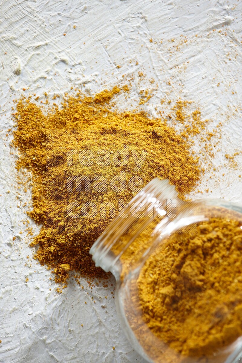 A flipped glass spice jar full of turmeric powder and powder fell out it on textured white flooring