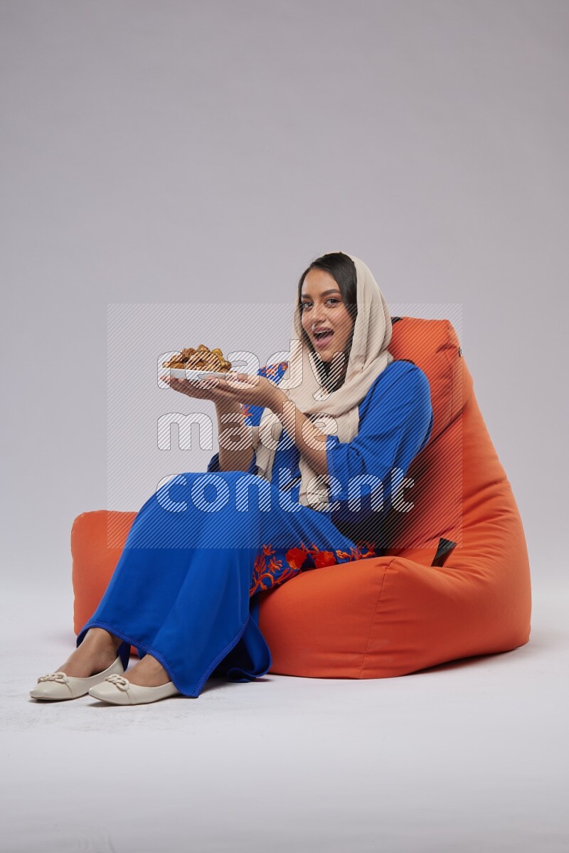 A Woman sitting on an orange beanbag wearing Jalabeya holding a plate of dates