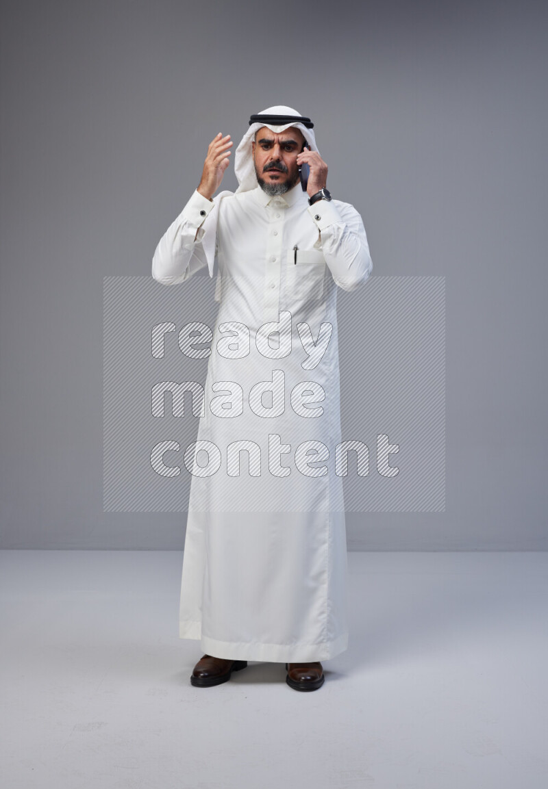 Saudi man Wearing Thob and white Shomag standing talking on phone on Gray background