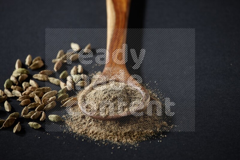 A wooden ladle full of cardamom powder and cardamom seeds beside it on black flooring