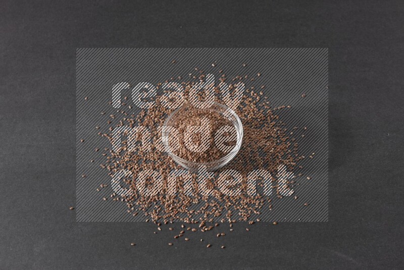 A glass bowl full of flaxseeds surrounded by the seeds on a black flooring