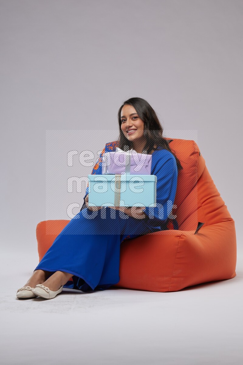 A woman sitting on an orange beanbag wearing Jalabeya holding a gift box