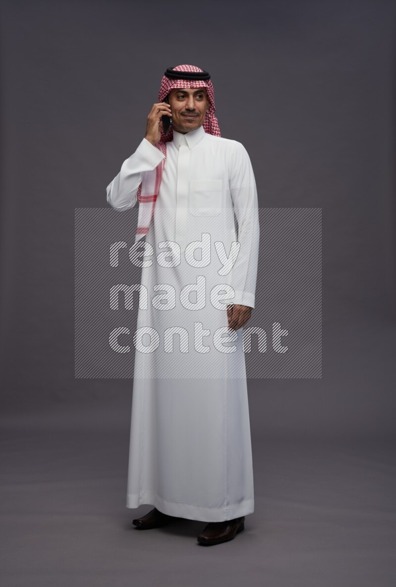 Saudi man wearing thob and shomag standing talking on phone on gray background