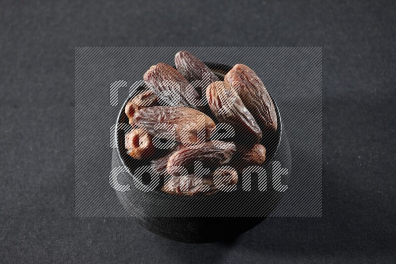 A black pottery bowl full of dried dates on a black background in different angles