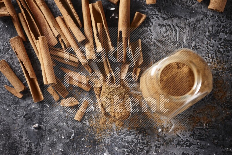 Herbal glass jar and a metal spoon full of cinnamon powder surrounded by cinnamon sticks on textured black background